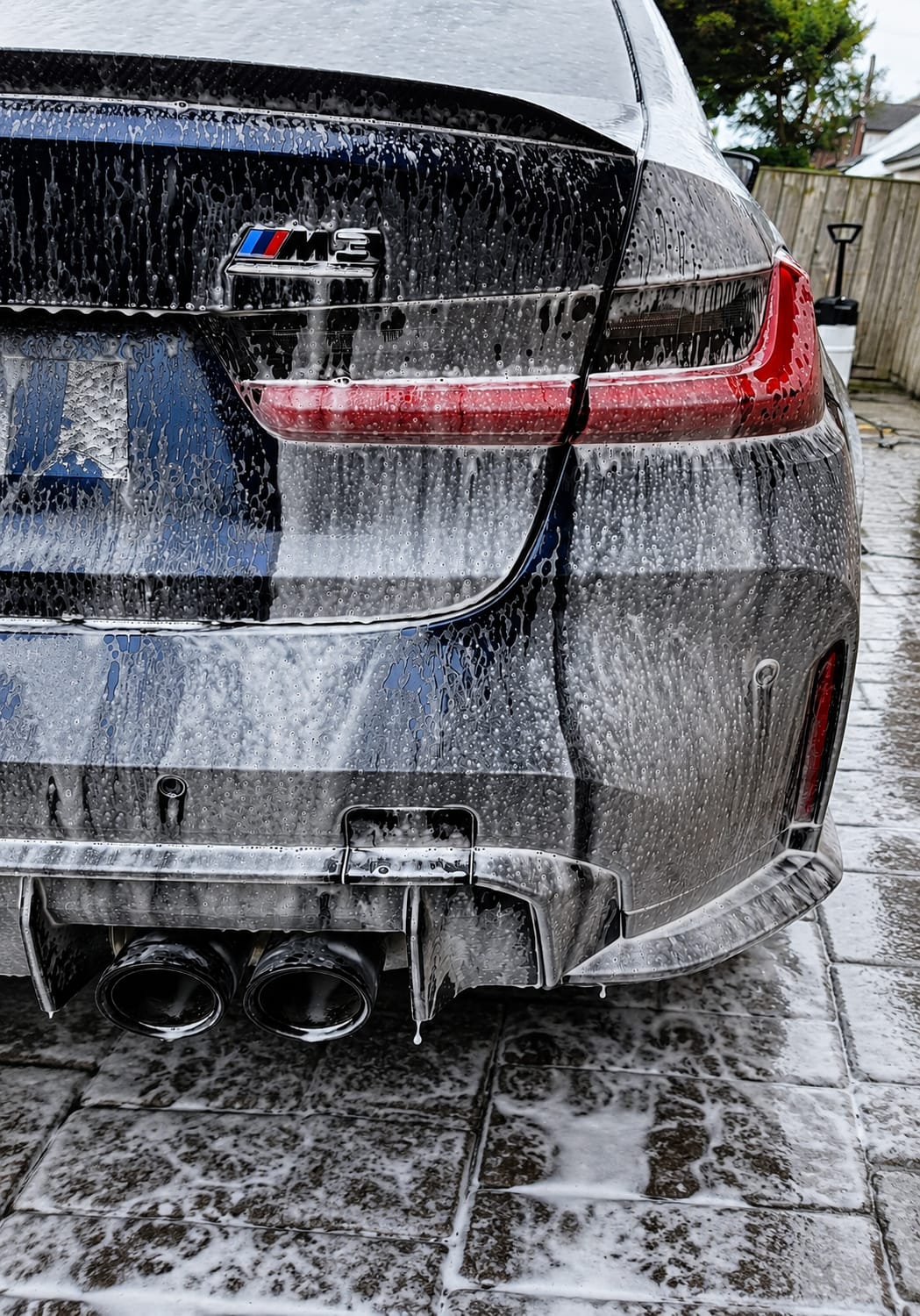 Rear of a dark BMW M3 covered in snow foam suds during a maintenance wash